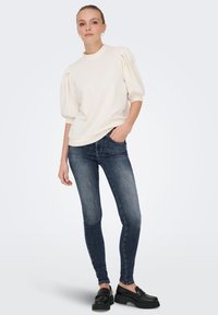 Pulover krem boje s naduvanim rukavima, uparen s tamnoplavim uskim trapericama i crnim mokasinkama. Glatka tekstura, casual kroj i minimalan dizajn.