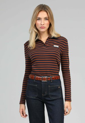 Polo à manches longues avec des rayures horizontales bleu marine et rouille, col à trois boutons, et un petit écusson portant l'inscription "1985". Jean en denim foncé avec poches.