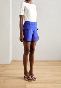 White short-sleeve top paired with blue shorts featuring two front pockets and gold hardware accents. Beige strappy heeled sandals.