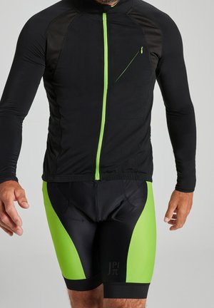 Homme portant une veste de cyclisme noire et vert néon avec fermeture éclair frontale et un cuissard de cyclisme rembourré assorti.
