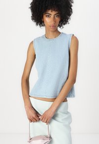 Svetlo modra denim brezrokavnik s krožno všečnico, ukrivljenim robom in vidnim šivom. Model drži majhno roza torbico.