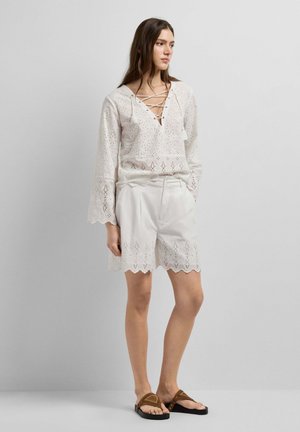 Jeune femme portant un chemisier en dentelle ajourée blanche et un short assorti à bords ondulés, debout devant un fond clair uni.