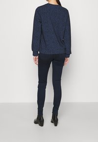 Marineblauw langemouwen shirt met een gestructureerde luipaardprint, gecombineerd met donkere skinny jeans en zwarte enkellaarsjes met hak. Effen achtergrond.