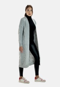 Lång ljusgrå stickad kappa med kabelstickade mönster, öppen front, ribbade ärmmuddar, bärs över en svart polotröja och glänsande svarta leggings, tillsammans med rosa sneakers.