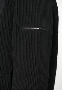 Crni pleteni pulover s rebrastom teksturom, prilagođenim dizajnom i džepom sa patentnim zatvaračem na lijevom rukavu s brendiranim detaljem.