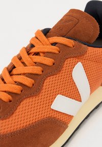 Oransje sneakers med mesh- og semsket skinntekstur, med hvit logo-detalj, runde snørebånd og en kontrastfarge i mørkeblå interiør.