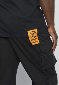 Svart T-skjorte med en oransje etikett som viser "240gsm" og merkevaren sin logo. Stoffet ser glatt ut, mens cargo-buksene har sidelommer.