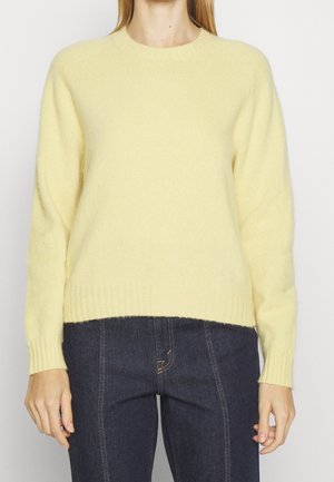 Personne portant un pull doux jaune pâle à manches longues et un jean bleu foncé avec surpiqûres contrastantes, vue de face du cou aux hanches.