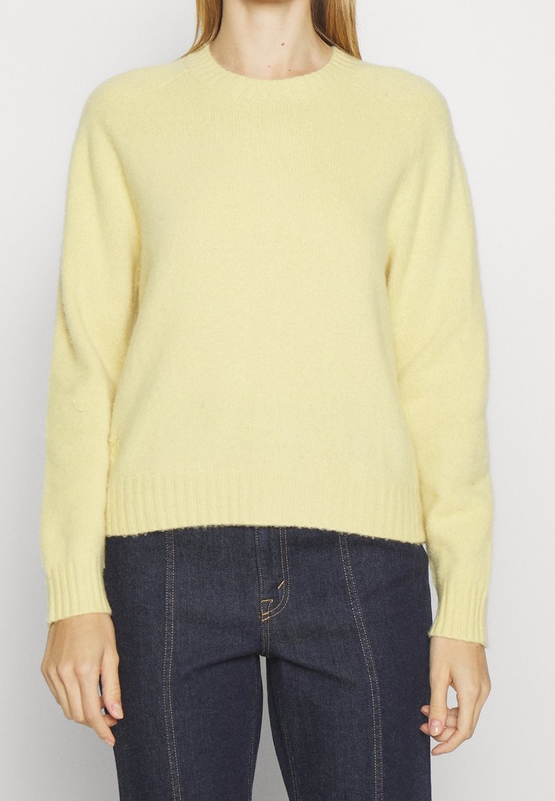 Personne portant un pull doux jaune pâle à manches longues et un jean bleu foncé avec surpiqûres contrastantes, vue de face du cou aux hanches.