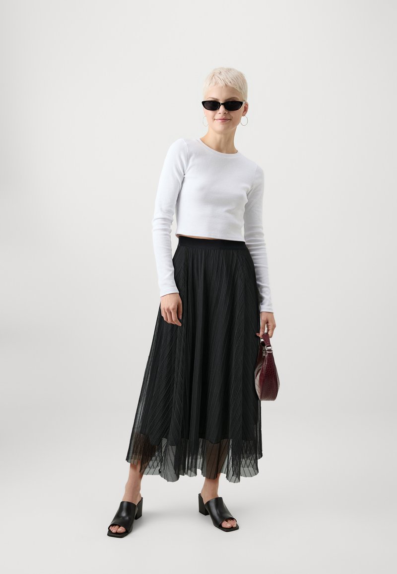 Schwarzer plissierter Maxi-Rock, weißes figurschmeichelndes Langarmoberteil, schwarze offen-toe Blockabsatz-Mules und burgunderfarbene Handtasche. Eine Sonnenbrille rundet den Look ab.