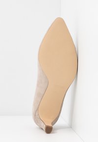 Chaussure à talon haut en daim beige avec un bout pointu et des détails délicats découpés au laser. Elle possède une semelle lisse et un petit talon.