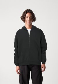 Black zip-up hoodie with a drawstring hood, made of soft fabric, featuring large front pockets and ribbed cuffs. Model wears it over a white shirt.