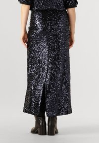 Zwarte paillettenrok met een lange, slanke vorm, voorzien van een zij-split. Gecombineerd met donkere, kniehoge laarzen met blokhak.