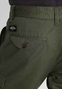 Olivgröna cargobyxor i bomullstyg med en bakficka med knappstängning och en Schott NYC-logotyp på midjebandet.