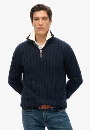 Suéter de punto azul marino con patrón de trenzas, diseño de media cremallera y un cuello blanco por debajo, combinado con jeans azules y un cinturón marrón.