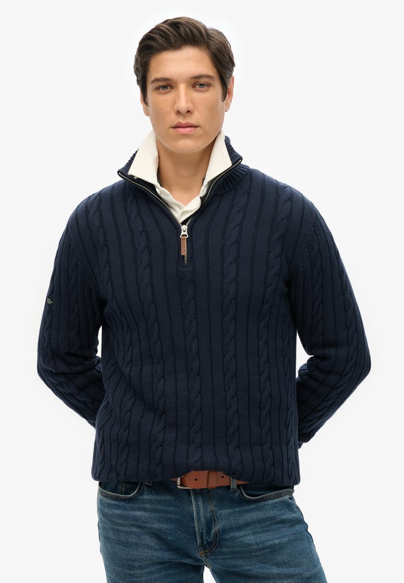 Marineblauwe gebreide trui met een kabelpatroon, ontwerp met halve rits en een witte kraag eronder, gedragen met blauwe jeans en een bruine riem.