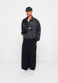 Svart mønstrete vindjakke med en lomme foran, i kombinasjon med løse svarte bukser og hvite sneakers. Modellen holder også en svart veske.