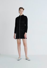 Svart zip-up klänning med lång ärm, tillverkad av texturerat tyg. Har en silverfärgad dragkedja och en strömlinjeformad silhuett, i kombination med silverfärgade platta skor.