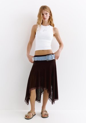 Jeune femme portant un haut blanc sans manches avec un logo dauphin, une jupe noire asymétrique, une large ceinture bleue et des sandales beiges, debout contre un mur blanc.