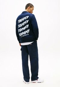 Marine sweatshirt met herhaalde witte "TOMMY" tekst op de achterkant; gecombineerd met marine cargobroek en witte sneakers. Moderne, casual streetwear stijl.