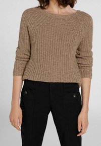 Maglione beige a maglia con una texture a costine, scollatura ampia e maniche arrotolate, indossato su pantaloni neri con tasche con bottoni.