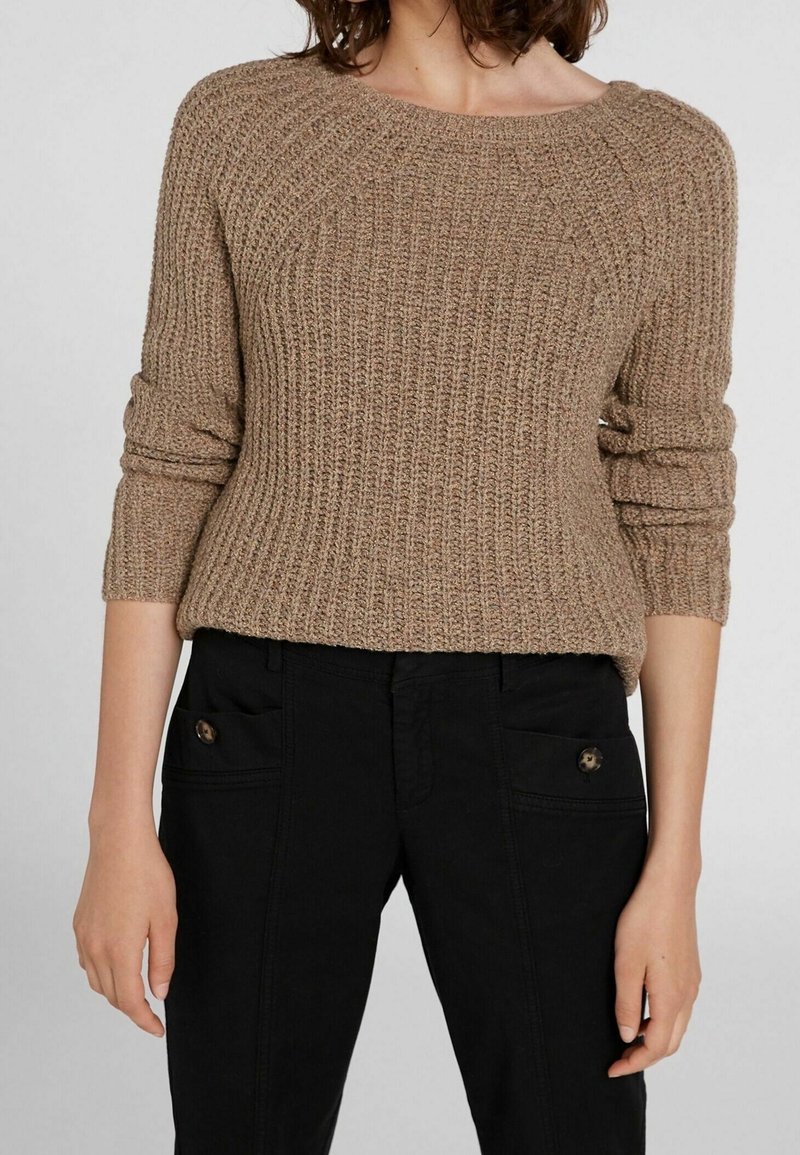 Maglione beige a maglia con una texture a costine, scollatura ampia e maniche arrotolate, indossato su pantaloni neri con tasche con bottoni.