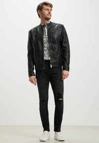 Giacca in pelle nera con zip frontale, dettagli sulle spalle, abbinata a una camicia a fantasia e jeans neri sdruciti. Scarpe da ginnastica bianche completano il look.