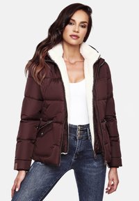 Brown puffer jacket with a faux fur-lined hood, zippered front, side pockets, and a quilted design. Worn over a white top and blue jeans.
