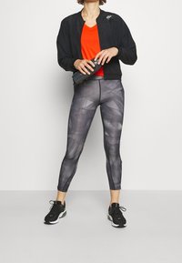 Giacca bomber nera, maglietta arancione a scollo a V, leggings grigi con motivo geometrico e scarpe da ginnastica nere. Tiene una bottiglia d'acqua.