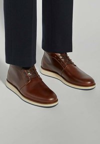 Brown leather ankle boots with a rounded toe, laces, and a white rubber sole. The design features a smooth texture and minimal stitching accents.