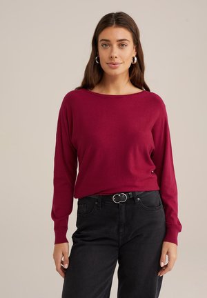 Maglione borgogna a maniche lunghe con scollatura ampia, texture morbida e vestibilità comoda, abbinato a jeans neri a vita alta e una cintura con fibbia argentata.
