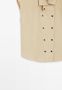 Beige sleeveless shirt with wide collar and double row of black snap buttons down the front.