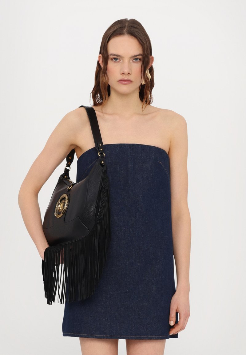 Zwarte leren handtas met franjes en metalen accenten; gedragen op de schouder. De tas heeft een gestructureerd oppervlak en een ronde vorm.