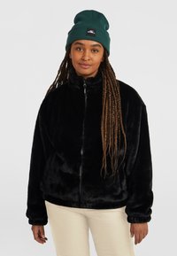 Zwarte faux fur jas met een hoge kraag en ritssluiting; heeft een relaxte pasvorm en elastische manchetten. Model draagt een groene beanie.