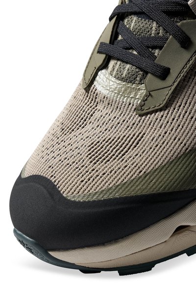 Gros plan sur l'avant d'une basket tricotée beige et olive avec des lacets noirs, un embout en caoutchouc et des détails de semelle texturée.