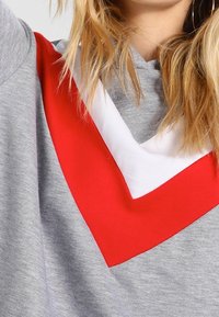 Szara bluza z czerwono-białym wzorem w kształcie litery V na klatce piersiowej. Miękka w dotyku, luźnego kroju, z kapturem i szerokimi rękawami.