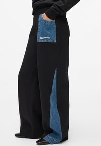 Pantalones de pierna ancha negros que presentan un gran bolsillo de parche de denim azul y detalles laterales. La tela tiene un acabado suave con una textura contrastante.