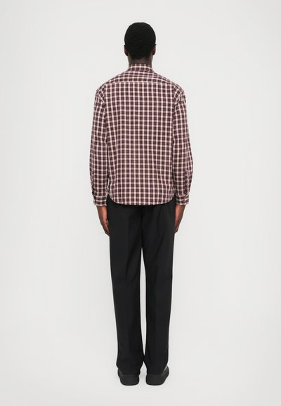 Uomo in piedi di spalle, che indossa una camicia a quadri bordeaux e bianca, pantaloni neri e scarpe nere, su uno sfondo bianco uniforme.
