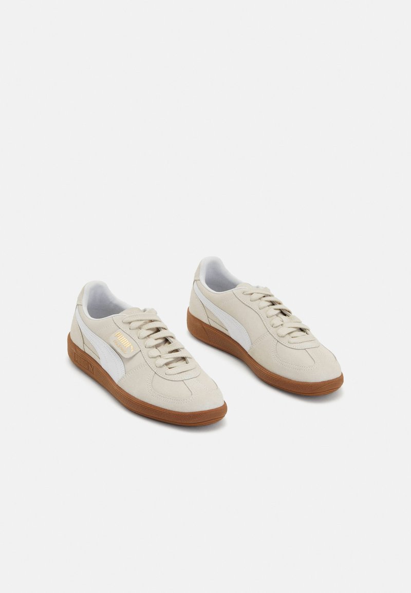 Beige Wildleder-Sneaker mit weißen Lederakzenten und braunen Gummisohlen. Sie verfügen über eine runde Zehenpartie, Schnürsenkel und ein goldenes Logo an der Seite.