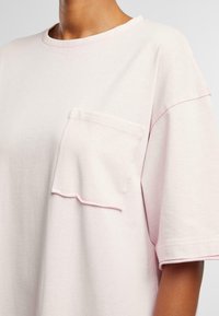 Tricou oversized roz deschis din bumbac moale, cu un buzunar unic cu margine zimțată și cusături contrastante de-a lungul gâtului.