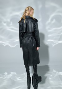 Černý kožený trenčkot s páskem v pase, detaily na knoflících a splývavým střihem, stylizovaný s ladícími kozačkami nad kolena.