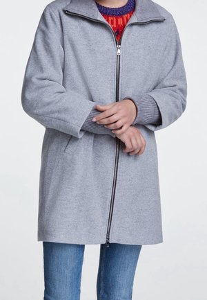 Person iført en lysegrå jakke med lynlås over en rød og lilla sweater, med hænderne foldet og blå jeans synlige under jakken.