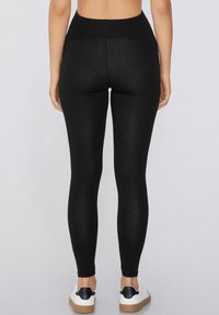 Leggings negros con una cintura alta, hechos de un material elástico. Textura suave sin patrones visibles. Llevados con zapatillas blancas.