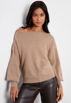 Kvinde med langt mørkt hår iført beige off-shoulder sweater med ballonermer og mørkebrune læderbukser, stående mod en hvid væg.