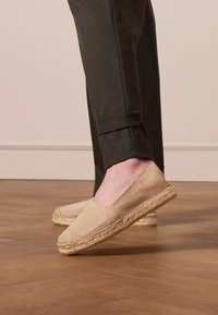 Beige espadrilles met een canvas bovenkant en jute zool, met een eenvoudig ontwerp en een lage profiel, gedragen met donkere broek op een houten vloer.