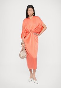 Koralinė satino suknelė su mazgo detalėmis ties juosmeniu, laisvai krentančiomis rankovėmis ir midi ilgio. Papildyta smėlio spalvos rankine ir baltais batais.