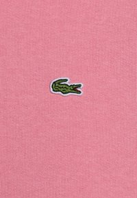 Zeleni izvezeni krokodil logo na ružičastoj rebrastoj tkanini.