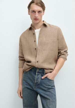 Junger Mann mit blondem Haar, der ein beiges Leinenhemd über einem weißen Oberteil und blauer Jeans trägt, stehend mit einer Hand in der Tasche.