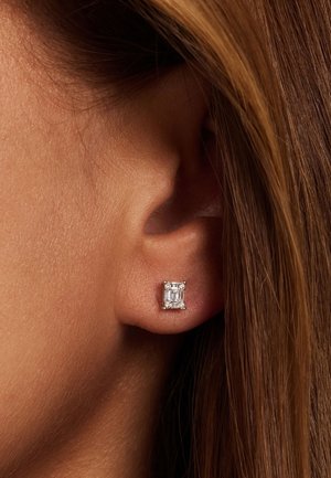 Boucle d'oreille en diamant taillé carré, montée sur un fond en argent poli, mettant en avant sa clarté et son éclat, portée sur l'oreille d'un modèle.