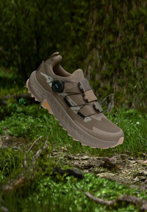 Scarpa da trail running marrone con lacci regolabili a quadrante che fluttua sopra un pavimento forestale roccioso e erboso.